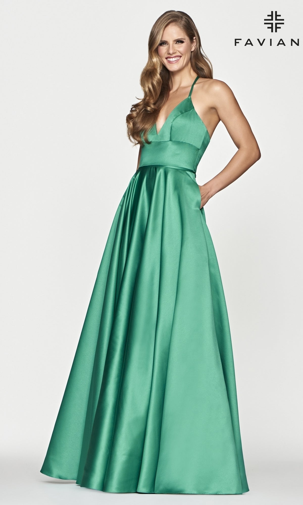 A woman wears the Corset-Back A-Line Long Satin Prom Dress by Faviana—sleeveless, emerald green satin with a deep V-neckline and pockets—standing against a plain white background.